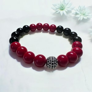 May include: A beaded bracelet featuring alternating red and black beads. A central bead is encrusted with black stones. The bracelet is displayed on a white surface, with white flowers in the background. This jewelry piece is suitable for everyday wear.