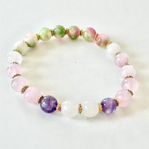 May include: A beaded bracelet featuring a variety of colorful gemstones. The bracelet includes pink, white, green, and purple beads, separated by gold-colored spacers. The gemstones are round and vary in size, creating a unique and eye-catching accessory.