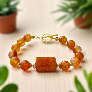 May include: A beaded bracelet featuring orange and gold-colored beads. The bracelet has a rectangular orange center stone and a gold-colored clasp. The beads are a mix of round and irregular shapes, creating a unique texture.