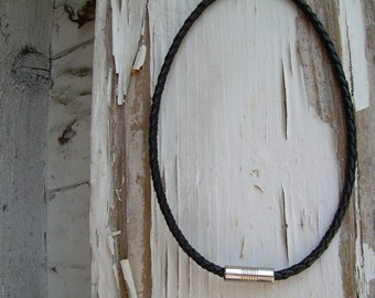 Puede incluir: Un collar de cuero negro con un cierre magnético plateado. El collar está trenzado y tiene un diseño sencillo.