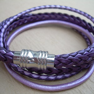 May include: A multi-strand wrap bracelet in shades of purple. The bracelet features braided leather strands and a smooth, solid strand. A silver-colored magnetic clasp secures the bracelet. The bracelet is designed to wrap multiple times around the wrist.