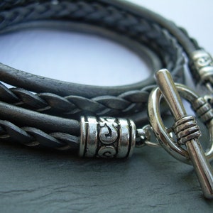 May include: A braided leather bracelet with a silver toggle clasp and decorative bead. The bracelet is made of three strands of leather that are braided together.