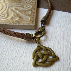 May include: A brown braided leather cord necklace featuring an antique brass-toned metal triquetra pendant. The necklace includes a matching brass-toned lobster claw clasp and decorative metal end caps with intricate embossed patterns.