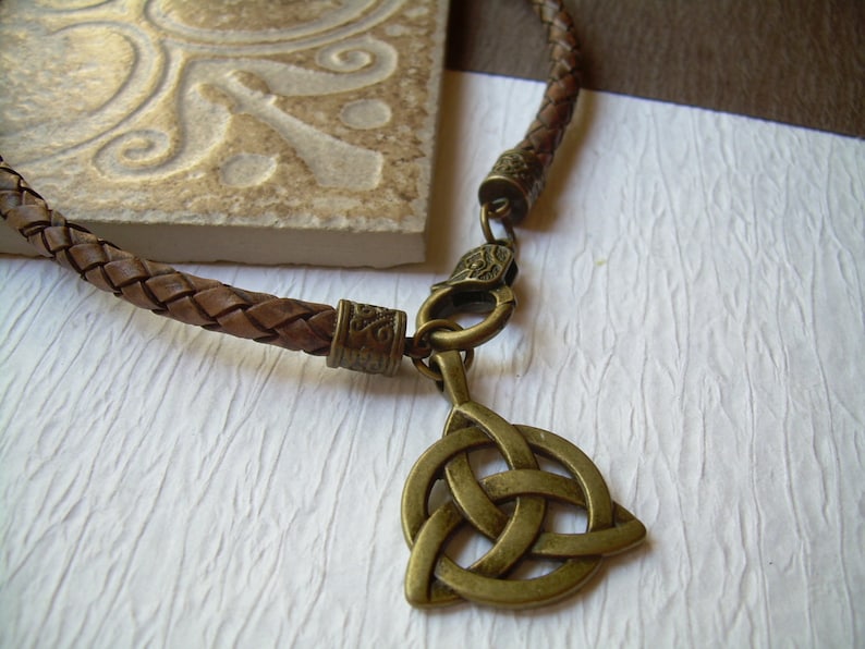 May include: A brown braided leather cord necklace featuring an antique brass-toned metal triquetra pendant. The necklace includes a matching brass-toned lobster claw clasp and decorative metal end caps with intricate embossed patterns.