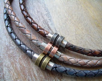 May include: Close-up of three braided leather necklaces in shades of brown, grey, and blue. Each necklace features a metal clasp with a different finish: silver, copper, and brass. The necklaces are displayed on a wooden surface.