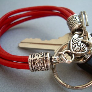 Puede incluir: Un brazalete de cuero rojo con un dije de corazón plateado y un llavero plateado.