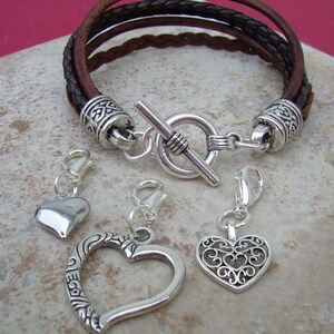 May include: A brown leather bracelet with a silver toggle clasp and three silver heart charms. The charms are a solid heart, a heart with an openwork design, and a heart with a filigree design.