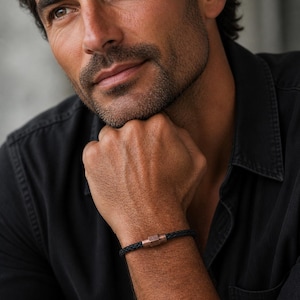 May include: Black braided leather bracelet with a copper-colored clasp. The bracelet is worn on a wrist, showcasing the design and texture of the leather and metal. The bracelet appears to be a stylish accessory.