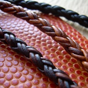 K&ouml;nnte beinhalten: Zwei geflochtene Lederb&auml;nder, eines braun und eines schwarz, liegen auf einer strukturierten orangen Oberfl&auml;che. Das braune Band hat einen silbernen Metallanh&auml;nger am Ende.