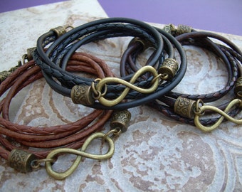 Op de afbeelding: Drie leren armbanden met oneindigheidssymbool bedels. De armbanden zijn bruin, zwart en een combinatie van bruin en zwart. De bedels zijn gemaakt van messing en hebben een versierd ontwerp.