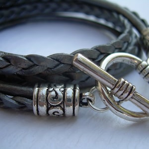 May include: A braided leather bracelet with a silver toggle clasp. The bracelet is made of three strands of leather that are braided together. The clasp is a simple, round design with a silver bar that slides through a loop.