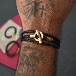Könnte beinhalten: Ein schwarzes Lederarmband mit einem goldfarbenen Verschluss. Das Armband wickelt sich mehrmals um das Handgelenk. Weitere Armbänder sind im Hintergrund sichtbar. Das Armband ist ein stilvolles Accessoire.