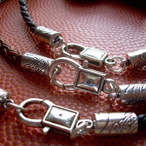 K&ouml;nnte beinhalten: Zwei Lederarmb&auml;nder mit silbernen Verschl&uuml;ssen und dekorativen Perlen. Die Armb&auml;nder sind geflochten und haben eine braune und schwarze Farbgebung.