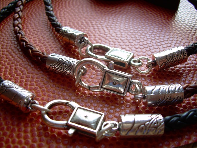 K&ouml;nnte beinhalten: Zwei Lederarmb&auml;nder mit silbernen Verschl&uuml;ssen und dekorativen Perlen. Die Armb&auml;nder sind geflochten und haben eine braune und schwarze Farbgebung.