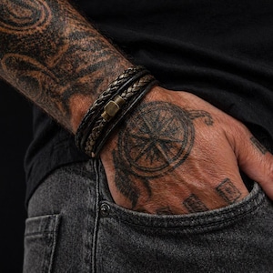 Puede incluir: Primer plano de tres pulseras de cuero marrón oscuro con un cierre de latón. Las pulseras se llevan en un brazo tatuado, con una camisa negra y vaqueros grises en el fondo.