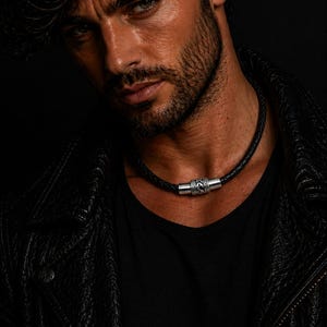 Puede incluir: Un collar gargantilla de cuero negro con cierre plateado. El collar se lleva alrededor del cuello. La imagen es un primer plano sobre un fondo oscuro. El collar es un accesorio de moda.