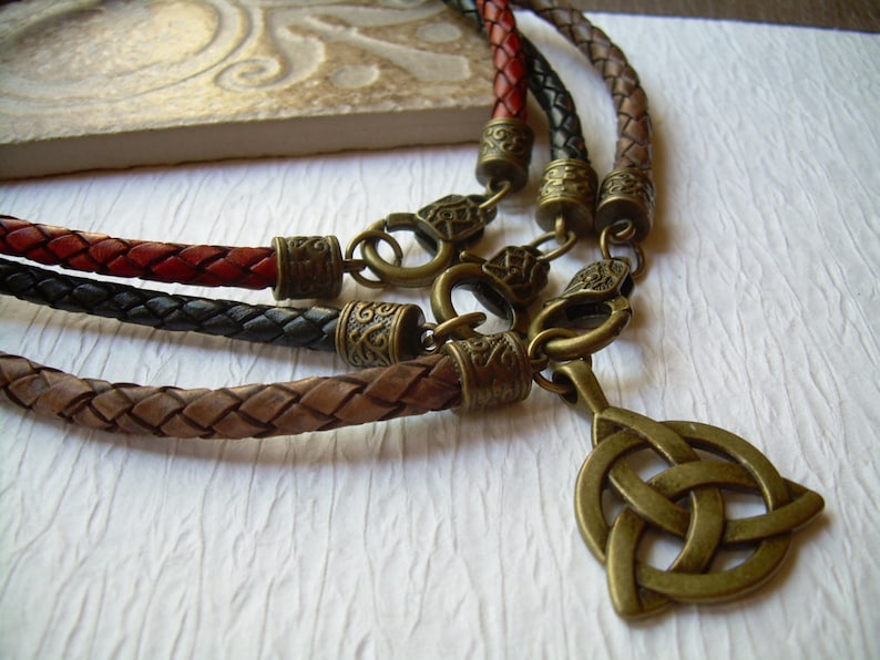 May include: Three braided leather cords in red, black, and brown, each featuring antique brass-toned metal end caps and lobster claw clasps. One cord includes a dangling antique brass-toned metal triquetra pendant with an intricate knot design.
