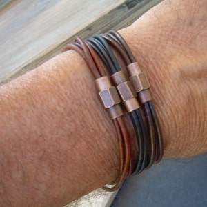 May include: A multi-layered leather bracelet with copper accents. The bracelet is made of brown and black leather and has a braided design.