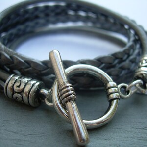 May include: A braided leather bracelet with a silver toggle clasp. The bracelet is made of three strands of leather that are braided together. The clasp is a simple toggle clasp with a silver ring and a silver bar.