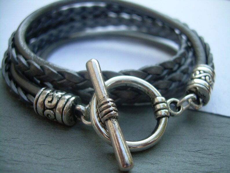 May include: A braided leather bracelet with a silver toggle clasp. The bracelet is made of three strands of leather that are braided together. The clasp is a simple toggle clasp with a silver ring and a silver bar.