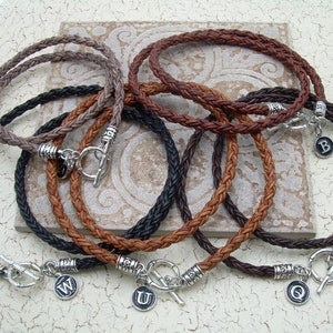Puede incluir: Seis pulseras de cuero trenzado con cierres de palanca plateados y dijes con letras. Las pulseras son de color marrón, negro y gris.