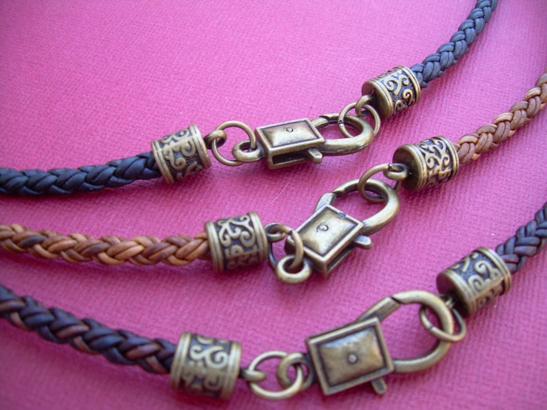 Op de afbeelding: Drie leren koordkettingen met decoratieve metalen sluitingen en kralen. De koorden zijn gevlochten in verschillende patronen en kleuren, waaronder bruin, zwart en beige.