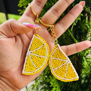 May include: Two lemon slice keyrings. Each keyring features a yellow beaded lemon slice with white beaded details, attached to a gold-coloured keyring. The keyrings are held up against a green leafy background.
