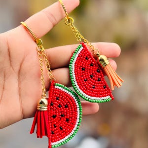Può includere: Due portachiavi a forma di fetta di anguria con disegni di perline rosse, verdi e bianche. Ogni portachiavi ha una catena dorata e un pon pon.