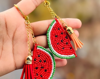 Könnte beinhalten: Zwei Schlüsselanhänger in Form von Wassermelonen-Scheiben mit roten, grünen und weißen Perlenmustern. Jeder Schlüsselanhänger hat eine goldene Kette und einen Quasten.