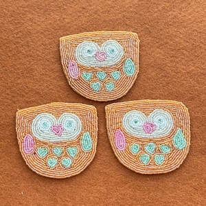 May include: Three beaded owl patches. Each patch features an owl design with orange, white, pink, and green beads. The owls have large eyes, heart-shaped details, and are set against a brown background. These patches are suitable for various craft projects.