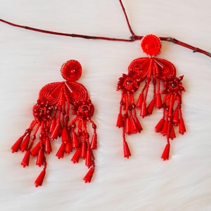 Może przedstawiać: Para czerwonych kolczyków z koralików z frędzlami. Kolczyki są wykonane z małych czerwonych koralików i mają dużą, okrągłą, czerwoną koralik na górze.