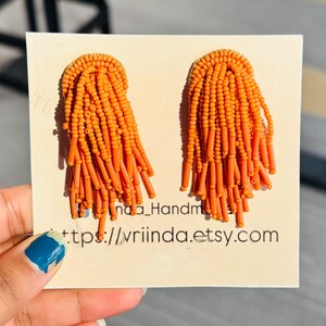 May include: A pair of orange beaded earrings. The earrings have a gold-colored base and are covered in small orange beads. The beads are arranged in a fringe design. The earrings are displayed on a cream-colored card with text.
