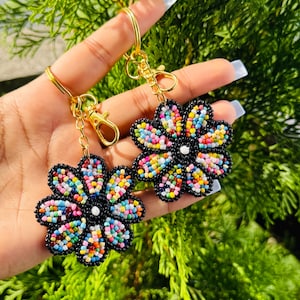 May include: Two beaded flower keychains. Each flower has a black outline and is filled with colourful beads. The keychains have gold-coloured hardware, including a clasp and a ring. The background is a blurred green.