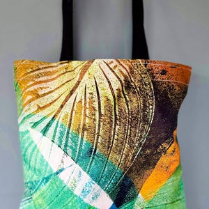 Könnte beinhalten: Eine farbenfrohe Tragetasche mit einem lebendigen abstrakten Design. Die Tasche hat eine Mischung aus grünen, blauen, orangefarbenen und braunen Farbtönen mit einem schwarzen Griff. Das Muster ähnelt einem stilisierten Blatt oder einer Feder.