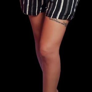 May include: A woman wearing black and white striped shorts with a black belt and black heels. She has a tattoo on her thigh.