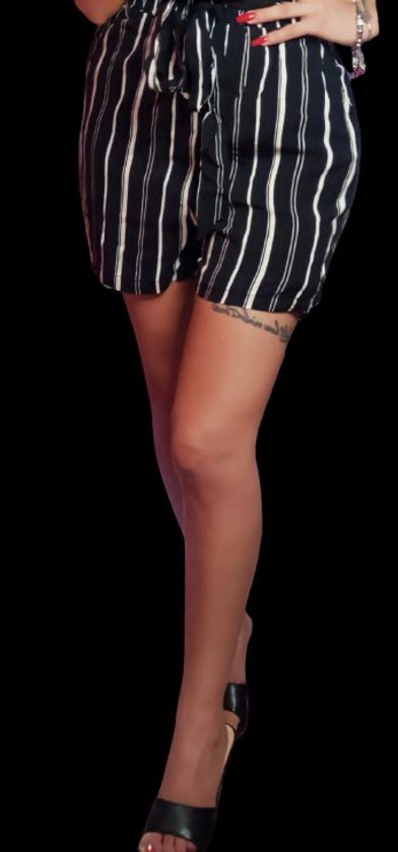 May include: A woman wearing black and white striped shorts with a black belt and black heels. She has a tattoo on her thigh.