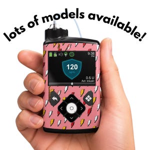 Può includere: Un dispositivo medico rosa con schermo nero che mostra parametri sanitari, tenuto in mano. Il dispositivo è decorato con un motivo di fulmini bianchi, gialli e neri. Lo schermo mostra 120 mg/dL e il testo "lots of models available!"