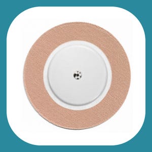 Puede incluir: Un parche adhesivo médico redondo de color beige con un pequeño agujero central.