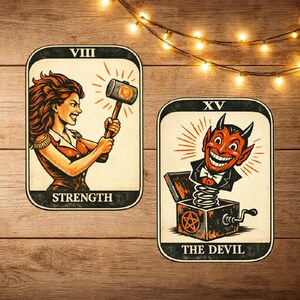 Puede incluir: Dos cartas de tarot sobre una superficie de madera. La carta "Fuerza" presenta a una mujer sosteniendo un martillo. La carta "Diablo" muestra a un diablo saliendo de una caja sorpresa. Ambas cartas tienen un estilo vintage ilustrado.