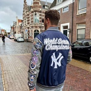 以下が含まれることがあります： 背面には「World Serie Champions」の文字と白い「NY」ロゴが入った、ネイビーブルーとグレーの野球ジャケット。袖はグレーで、複数のパッチが付いています。明るいブルーのジーンズを履いた人が着ています。
