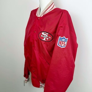 Könnte beinhalten: Rote Satin-Bomberjacke mit einem weiß-rot gestreiften Kragen und Bündchen. Die Jacke hat ein großes "SF"-Logo auf der Vorderseite und einen NFL-Patch am Ärmel.