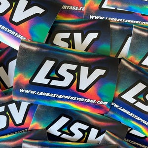 Puede incluir: Una pila de pegatinas holográficas iridiscentes con el texto "LSV" y el sitio web "www.laurastappersvintage.com" impresos en ellas.