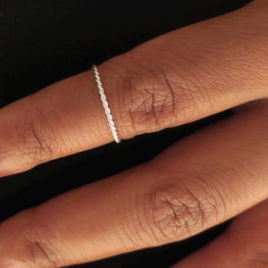 Könnte beinhalten: Ein dünner, silberfarbener Ring mit einer feinen, gedrehten Kordelstruktur. Das Band ist kreisförmig und behält über das gesamte Design eine gleichmäßige Breite bei, wobei es den Finger mit einer polierten, metallischen Oberfläche umschließt.