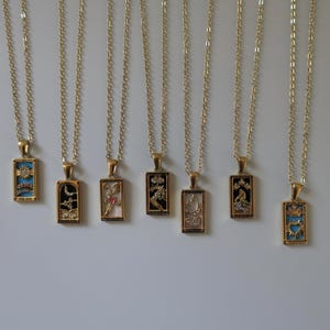 Könnte beinhalten: Sieben goldfarbene Halsketten mit rechteckigen Anhängern, die verschiedene vom Tarot inspirierte Motive zeigen. Jeder Anhänger weist einzigartige Darstellungen wie Himmelskörper, Herzen und Kronen auf, akzentuiert mit kleinen klaren Steinen und farbiger Emaille.