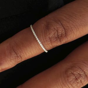 Könnte beinhalten: Ein dünner, silberfarbener Ring mit einer zarten, texturierten Oberfläche, die aus kleinen, sich wiederholenden quadratischen Gliedern besteht. Das Band hat ein metallisches Finish und ein schmales Profil, das den Finger mit einem gleichmäßigen Muster umschließt.