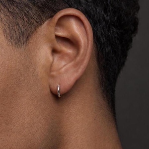 Könnte beinhalten: Ein kleiner, polierter Silber-Creolen-Ohrring, der im Ohrläppchen getragen wird. Der Ohrring hat ein schlichtes, kreisförmiges Design mit einer glatten, reflektierenden Metalloberfläche, die das Licht einfängt und einen subtilen Glanz auf der Haut erzeugt.
