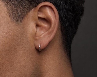 Könnte beinhalten: Ein kleiner, polierter Silber-Creolen-Ohrring, der im Ohrläppchen getragen wird. Der Ohrring hat ein schlichtes, kreisförmiges Design mit einer glatten, reflektierenden Metalloberfläche, die das Licht einfängt und einen subtilen Glanz auf der Haut erzeugt.