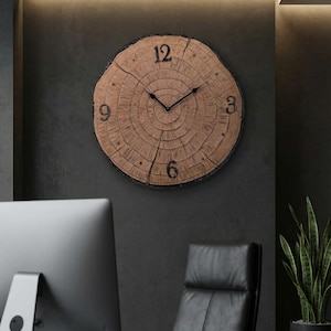 Puede incluir: Un reloj de pared grande y redondo con aspecto de veta de madera. La esfera del reloj presenta números negros en las posiciones 12, 3, 6 y 9, con manecillas negras. El reloj está montado en una pared gris oscuro, creando una estética rústica pero moderna.