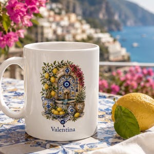 May include: A white ceramic mug featuring a colorful illustration of a stone fountain surrounded by lemon trees, potted plants, and blue and white patterned pottery. The design includes a blue evil eye symbol and the name Valentina printed in blue.