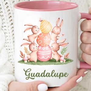 May include: White ceramic mug with a pink interior and handle. The mug features an Easter-themed design with bunnies, decorated eggs, and the name "Guadalupe" in green script. The design includes orange, pink, and yellow colors.